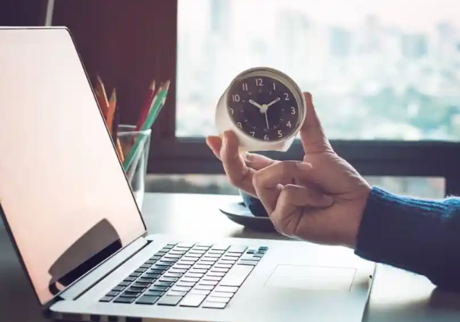 Hand holding a clock beside a laptop, representing the start of the workday
